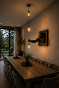 A long wooden dining table surrounded by gray upholstered chairs in a warmly lit dining room. A sculptural wall piece on the wall appears to be holding the hanging light fixture, and the scene includes pendant bulbs, candles, plants, and a large window looking out onto greenery.